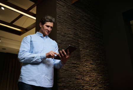 Confident pleasant Caucasian man, interior designer swipes websites on a digital tablet, standing in...
