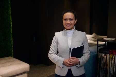 African pretty woman, sales manager, interior designer stands near stand with upholstery color fabric swatches in a furniture design showroom, holds a catalog and smiles looking confidently at cameraの写真素材