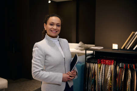 African American pleasant woman, sales manager, interior designer holds a catalog and smiles toothy smile looking at camera standing near a stand with color fabric samples in a furniture design studioの写真素材