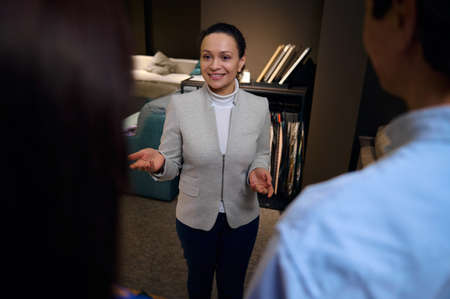 Adorable pleasant confident multiethnic female interior designer, sales manager in furniture design store talking with customers, standing against a stand with upholstery samples of fabrics for sofas.の写真素材