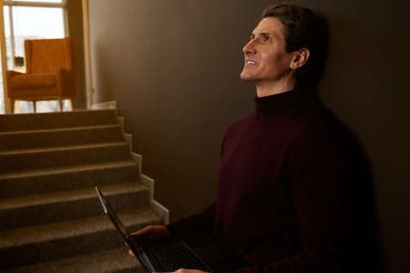 Side portrait of a handsome Caucasian man working remotely on laptop against dark brown wall background. Businessman, entrepreneur, copywriter, journalist freelance. Online work concept. Copy ad spaceの写真素材