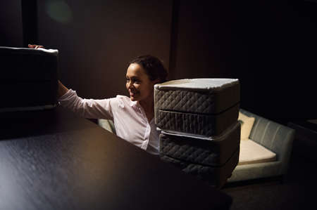Pretty African American woman sales assistant demonstrates samples of mattresses in a bed, mattress and pillow storeの写真素材