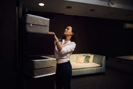 Charming dark-haired multi-ethnic woman, designer, retail consultant, sales manager, retailer, warehouse worker lays out samples of orthopedic mattresses on a standの写真素材