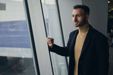 Portrait of charming handsome thoughtful Middle Eastern man, transit passenger standing at panoramic windows overlooking runway in the international airport departure terminal, waiting to board flightの写真素材