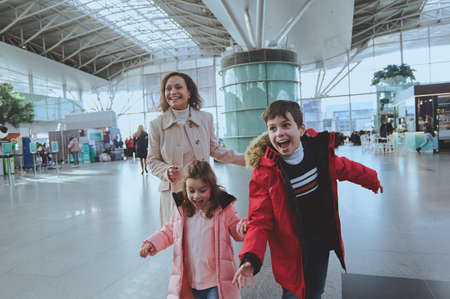 Happy multiethnic woman with her children running in airport arrivals terminal waiting room. Long-awaited family reuniting conceptの写真素材
