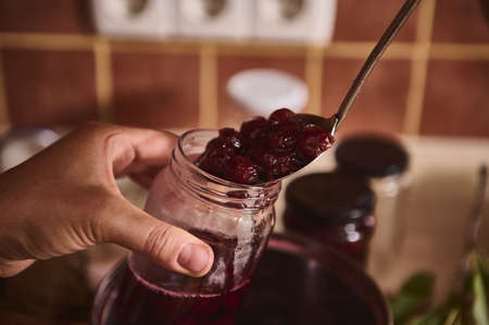 Close-up confectioner's hands holding kitchen spoon with cherry berries boiled in homemade delicious...