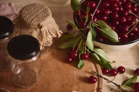 Close-up. Top view of sterilized glass jam jars with lids on a wooden board next to an enameled bowl of freshly picked cherries on a kitchen countertop. Preparation for conservationの写真素材