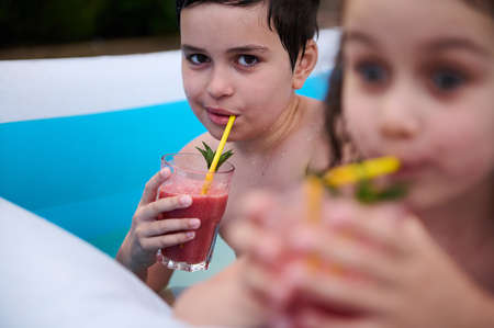 Handsome school age boy drinking freshly squeezed strawberry juice from a straw. Adorable baby girl on the blurred foreground. Kids having fun in an inflatable swimming pool at home gardenの写真素材