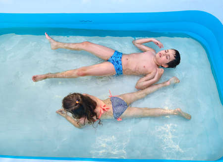 Top view of joyful Caucasian kids - boy and girl, brother and sister, enjoying their summer holidays, swimming in a inflatable swimming pool. Leisure activity, happy and carefree childhood conceptの写真素材