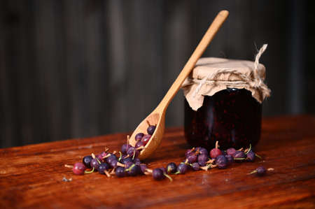 Still life. A glass jar of a homemade wild berry jam, wooden spoon and gooseberries on a rustic wood...