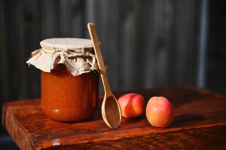 Close-up. Still life. A glass jar of homemade jam, with craft paper on the lid tied with rope, ripe ready-to-eat red sunny apricots and wooden spoon on rustic wood surface. Canning, sweet preserves.の写真素材