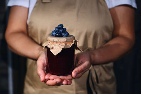 Canning, preserved food concept for winter season. Focus on a jar of blueberry jam with fresh berries on the craft lid, in the hands of a woman confectioner wearing a chef apron. Close-up.の写真素材