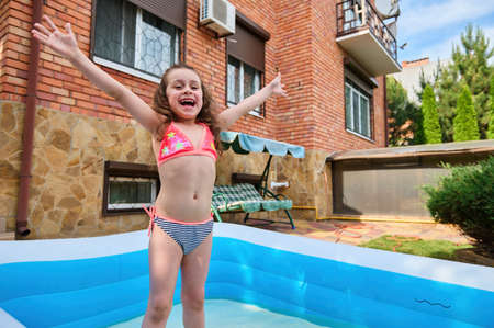 Cheerful Caucasian baby girl in a bright swimsuit having fun in the inflatable swimming pool in the door of a private house. Summer holidays concept. Happy childhood, leisure activityの写真素材