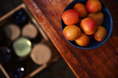 Soft focus on ripe, ready-to-eat apricots in a blue ceramic bowl on a wooden surface against a blurry wooden box with jam jars on the floor. Organic farm and canning conceptの写真素材