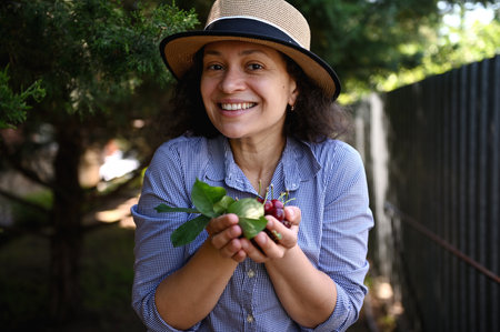 Pleasant multiethnic woman agronomist, farmer holds handful of harvested cherries and smiles sweetly, looking at camera.の写真素材