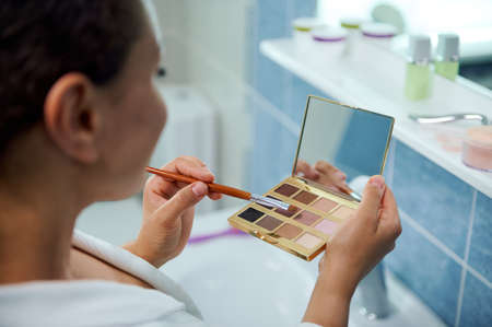 Rear view of a blurred woman holding eyeshadow palette with small cosmetic mirror and makeup brush, applying make-upの写真素材
