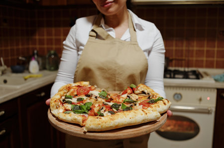 Homemade, freshly baked vegetarian pizza with addition basil herbs and mozzarella cheese on wooden board in the hands of charming woman, housewife in a chefs apron, standing at home kitchen interiorの写真素材
