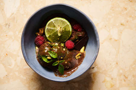Selective focus on a slice of juicy lime, garnishing a chocolate ice cream, topped with organic cocoa, raspberries and lemon basil leaves, melting in navy blue bowl. Flat lay Still lifeの写真素材