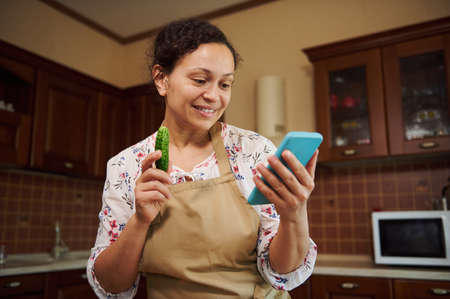 Pretty, natural beauty multiethnic woman, housewife wearing a chefs apron, looking through recipe of marinating and pickling seasonal vegetables at home kitchen. Close-up. Family traditionsの写真素材