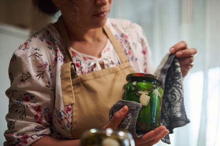 Cropped view of a middle aged woman, housewife in chefs apron, wrapping a glass jar with freshly canned marinated cucumbers in warm terry cloth kitchen towel. Pickling conceptの写真素材