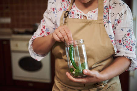 Details: Hands of a housewife in a kitchen apron, holding can and filling it with fresh chili pepper while pickling at home kitchen. Preparing homemade pickles for the winter. Food canning. Close-upの写真素材