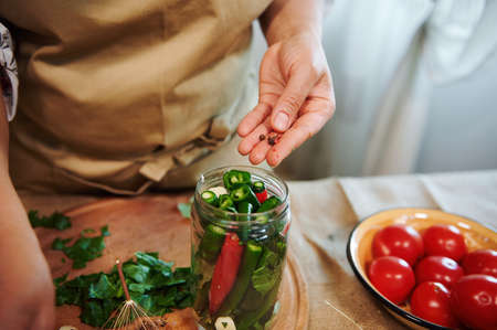 Details: Womans hand adding peppercorns in glass jar for canning, filled with stacked fresh chili peppers while making homemade spicy preserves at home kitchenの写真素材