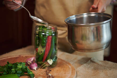Close-up. Housewife pours boiling marinade over chili peppers in jars. Chopped dill leaves and fresh garlic laying out on a wooden board. Pickling seasonal vegetable at home. Harvest conservation.の写真素材