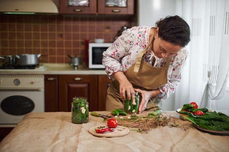 Pleasant multiethic woman, housewife, wearing a chefs apron preparing homemade pickled cucumbers for the winter in the home kitchen interior. Fresh ingredients on the table with linen tableclothの写真素材