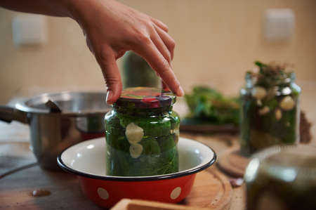 Details: Chefs hand putting a lid on a sterilized jar with cucumbers, pickled with fresh dill...