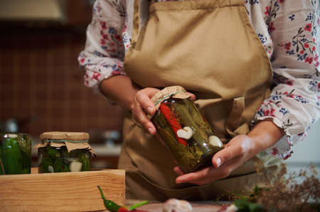 Close-up of the hands of a housewife in beige chefs apron, holding a glass jar of homemade pickled chili peppers. Marinating and preserving vegetables, healthy fermented food, winter natural productsの写真素材
