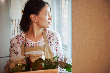 Pleasant multiethnic women, housewife in chefs apron thoughtfully looks through the window in the kitchen while holding a box with jars of pickled vegetables, preserved according to a homemade recipeの写真素材