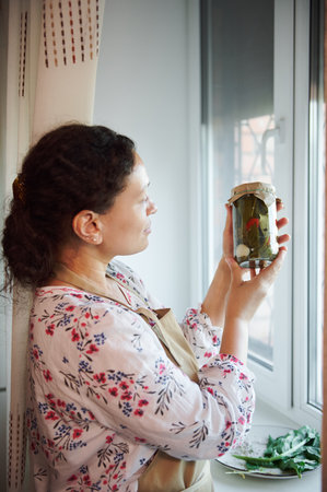 Portrait of serene mature woman, housewife in a chefs apron standing by window at home kitchen and looking at the jar with marinated chili peppers, canned according a traditional family recipeの写真素材