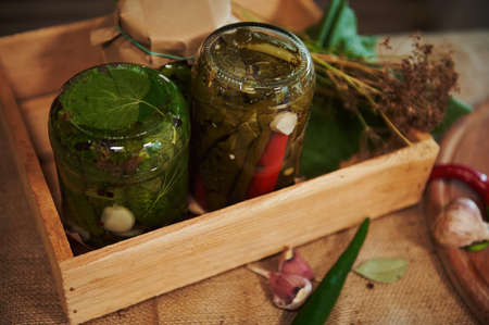 Still life. Jars of marinated chili peppers and pickled cucumbers, upside down on wooden board, with...