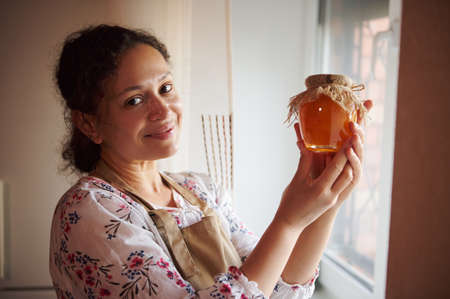 Hispanic pretty woman, pleasant housewife, stands by the window at home kitchen, smiles at the camera, presenting homemade peach jam, confiture, marmalade, canned according traditional family recipeの写真素材