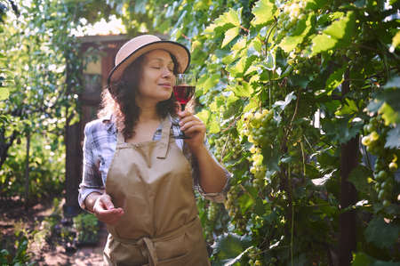 Charming curly haired woman in a straw hat tasting homemade wine, standing in the backyard near...