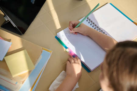 Top view with details of a schoolgirls hand holding pencil and learning the writing, sitting a desk in front of a digital gadget with an online broadcast of video lesson. Back to school. First gradeの写真素材