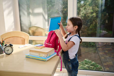 Side portrait. A little European girl with two pigtails, in a white T-shirt and blue denim overalls, stands at the table on the veranda of the house and puts her school supplies into a pink school bagの写真素材