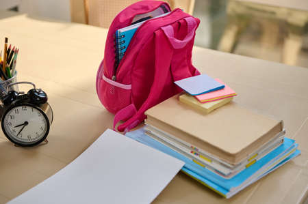 School supplies and stationery. Pink backpack and stacked textbooks near a black alarm clock on the table. Back to school concept. Copy space for advertising textの写真素材