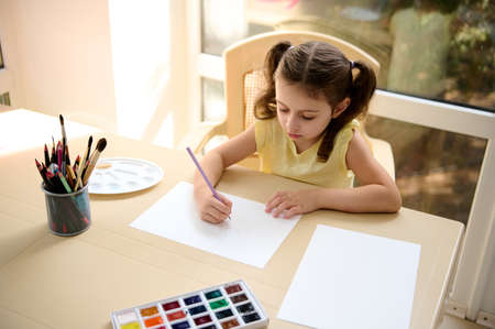 View from above of an inspired schoolgirl, talented artist painting an image on a white blank sheet...