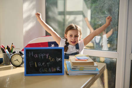 Adorable European little girl, a schoolgirl raising arms and clenching fists, enjoying the remote study at first grade in an online American school. Back to school. Knowledge. Education conceptの写真素材