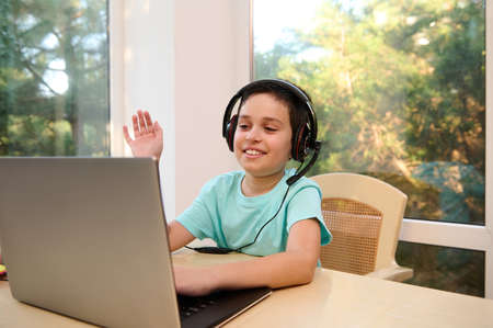 Adorable schoolboy, in headphones, waves with hand to the teacher while studying remotely from home,...