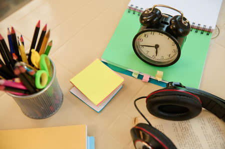 Still life. View from above with focus on a black alarm clock on open notepad with school supplies, audio headset and stationery on blurred foreground. Back to school. Education. Teachers day conceptの写真素材