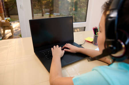 Rear viewof a teenage schoolboy in headphones sitting at laptop with copy space on blank monitor...