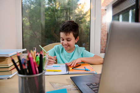 Front view of a cheerful happy schoolboy doing homework, learning geometry sitting at laptop during...