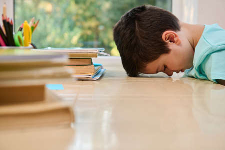 Tired bored schoolboy, primary student in online school, sitting at desk, leaning on the table with...