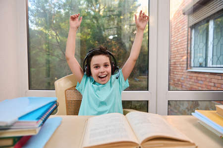 Happy schoolboy in headphones clenches his fists and raises his hands as a sign of victory and achievement of goals, sits at table with textbooks and smiles cutely at the camera. Online home schoolingの写真素材