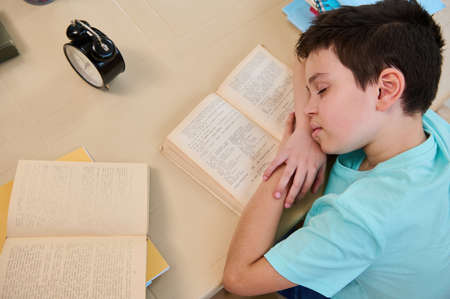 Overhead view of a primary school student, falling asleep on a textbook, feeling tired doing...