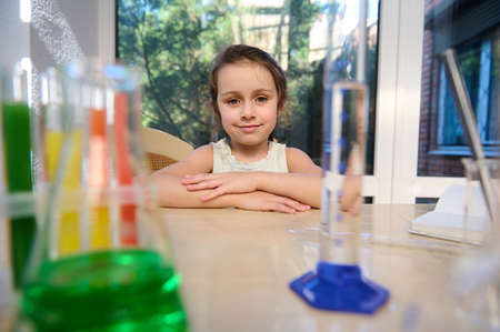 Adorable preschooler sitting at table with test tubes filled with colorful reagents, flasks,...