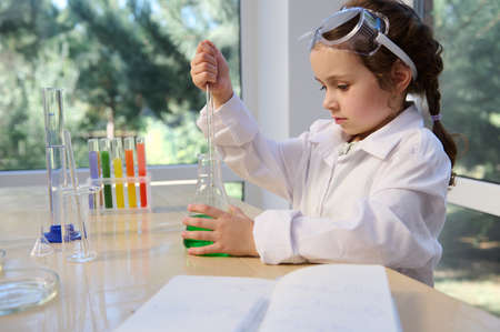 Little adorable elementary age girl using glass laboratory pipette, picking up a green solution from a flat-bottomed flask, while visiting a chemistry circle for little future chemists scientistsの写真素材
