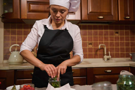 Beautiful female chef, skilled housewife in white cap and black apron filling the sterilized can for canning with fresh culinary herbs and spices, while pickling fresh organic harvest at home kitchenの写真素材